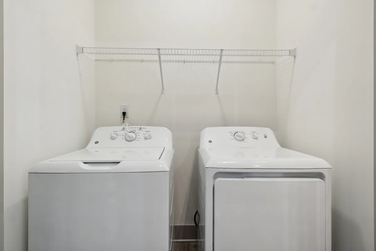 This image shows a dedicated laundry area equipped with a washing machine, drying machine, and a convenient shelf for organizing laundry supplies. The space is neat, practical, and well-lit, emphasizing modern convenience for day-to-day living.