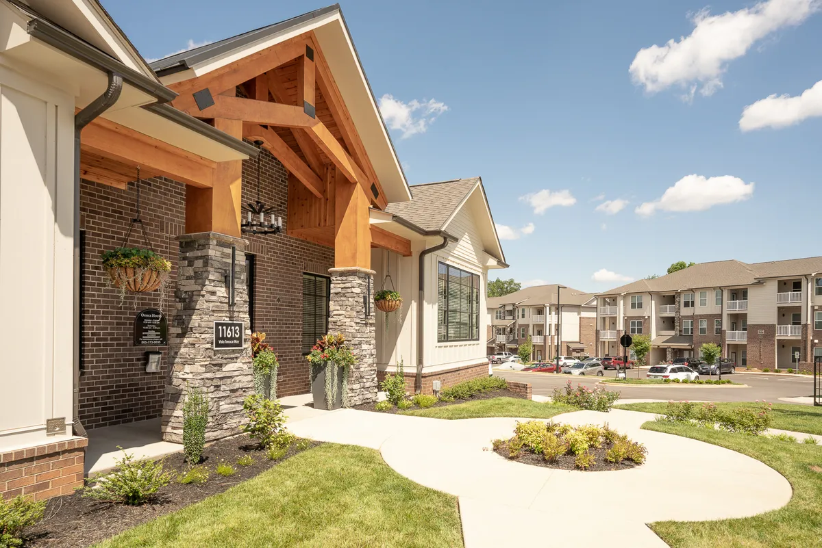 The inviting entrance features striking wooden beams, a stone facade, and landscaped garden accents. Large windows hint at the bright, open spaces inside, while the garden and gathering area suggest opportunities for community connections and relaxed socializing.