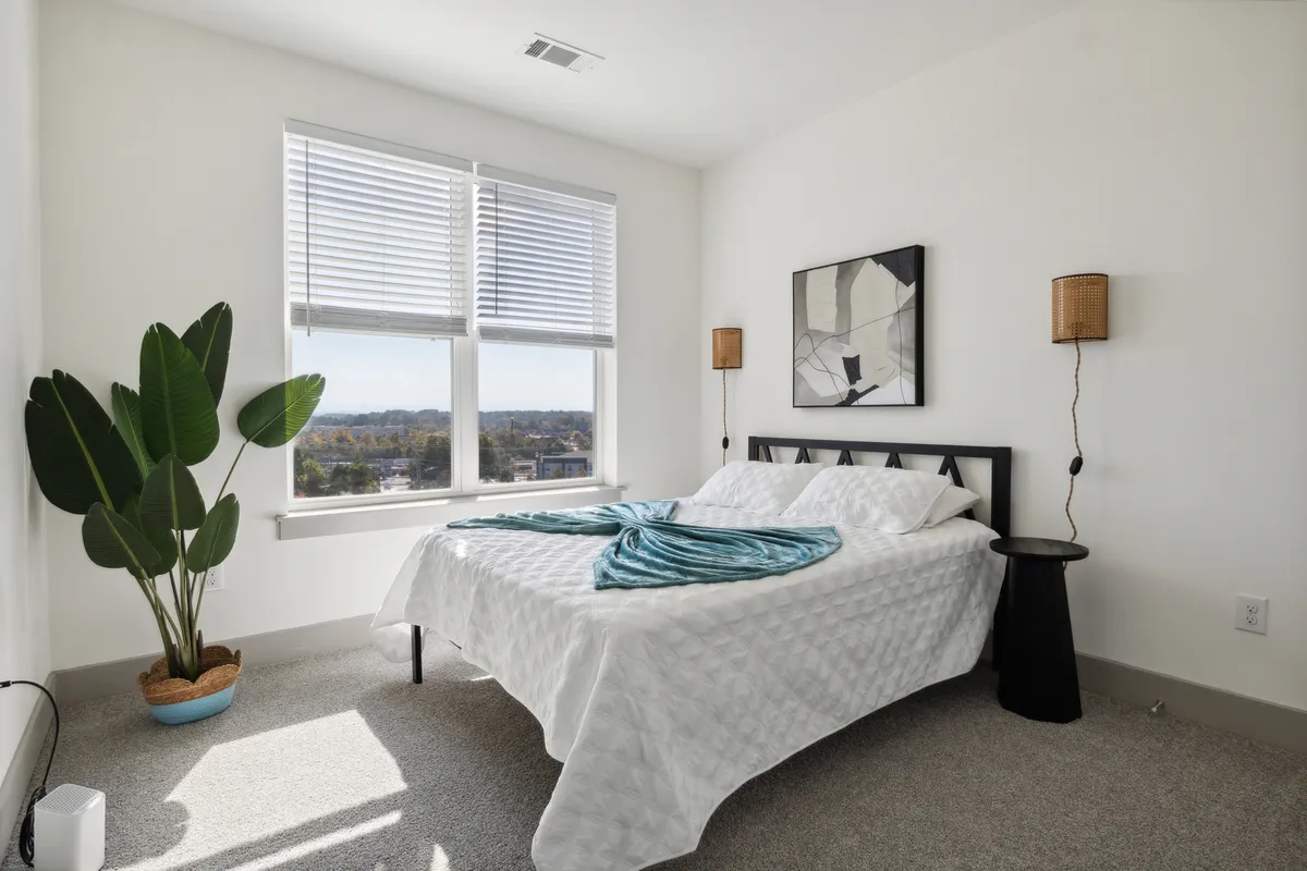 A tranquil bedroom with a bed dressed in a crisp white quilt, flanked by large windows, green plants, abstract artwork, and bedside lamps. The room is filled with natural light and offers a peaceful view, inviting relaxation and quiet moments.