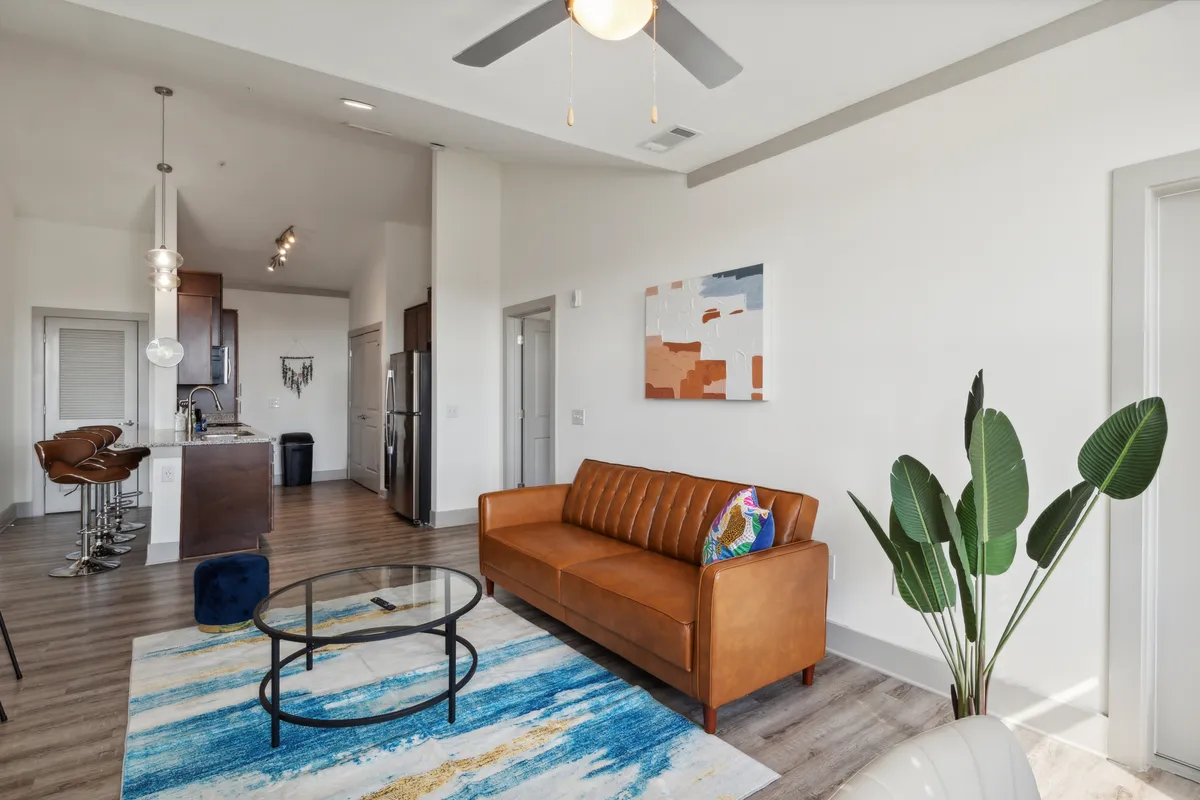 A stylish living room featuring a brown leather sofa, colorful artwork, a glass coffee table, and a potted plant. The space feels welcoming and comfortable, ideal for relaxing after a day out or entertaining guests.