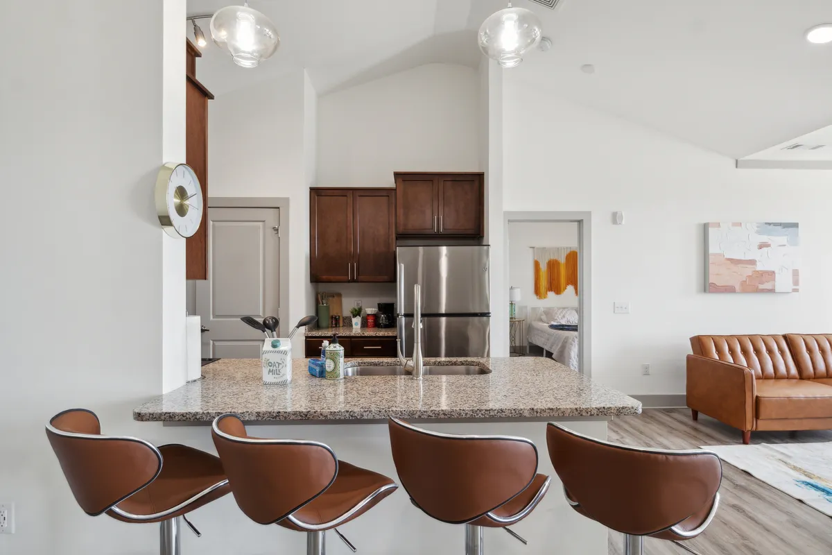 A welcoming view of the living area and kitchen, featuring a granite countertop with bar stools, modern appliances, and a cozy sofa. The space is designed for socializing and relaxing, with stylish decor and warm lighting creating an inviting atmosphere.