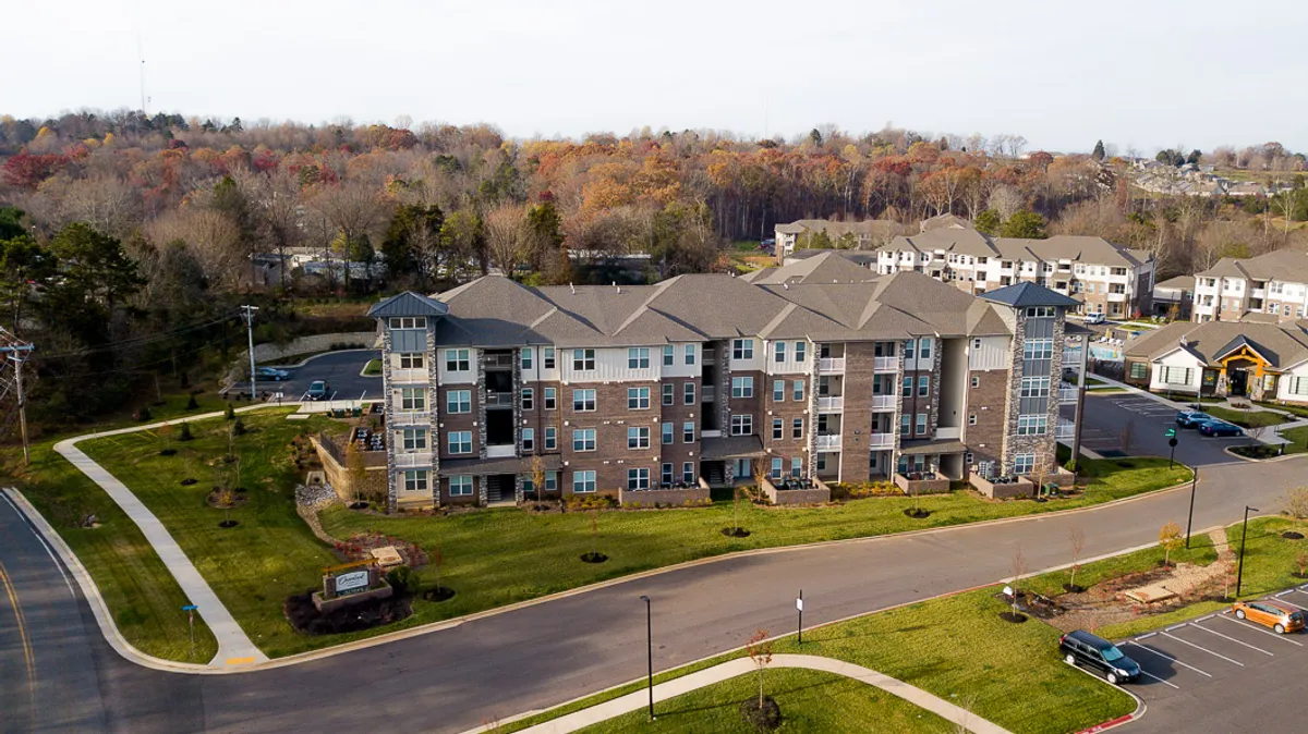 A sweeping view of the modern apartment complex is framed by meticulously landscaped green areas and a spacious parking lot. Residents can be seen strolling along the walkways, with the complex's welcoming architecture and lush outdoor spaces setting a vibrant, social tone.