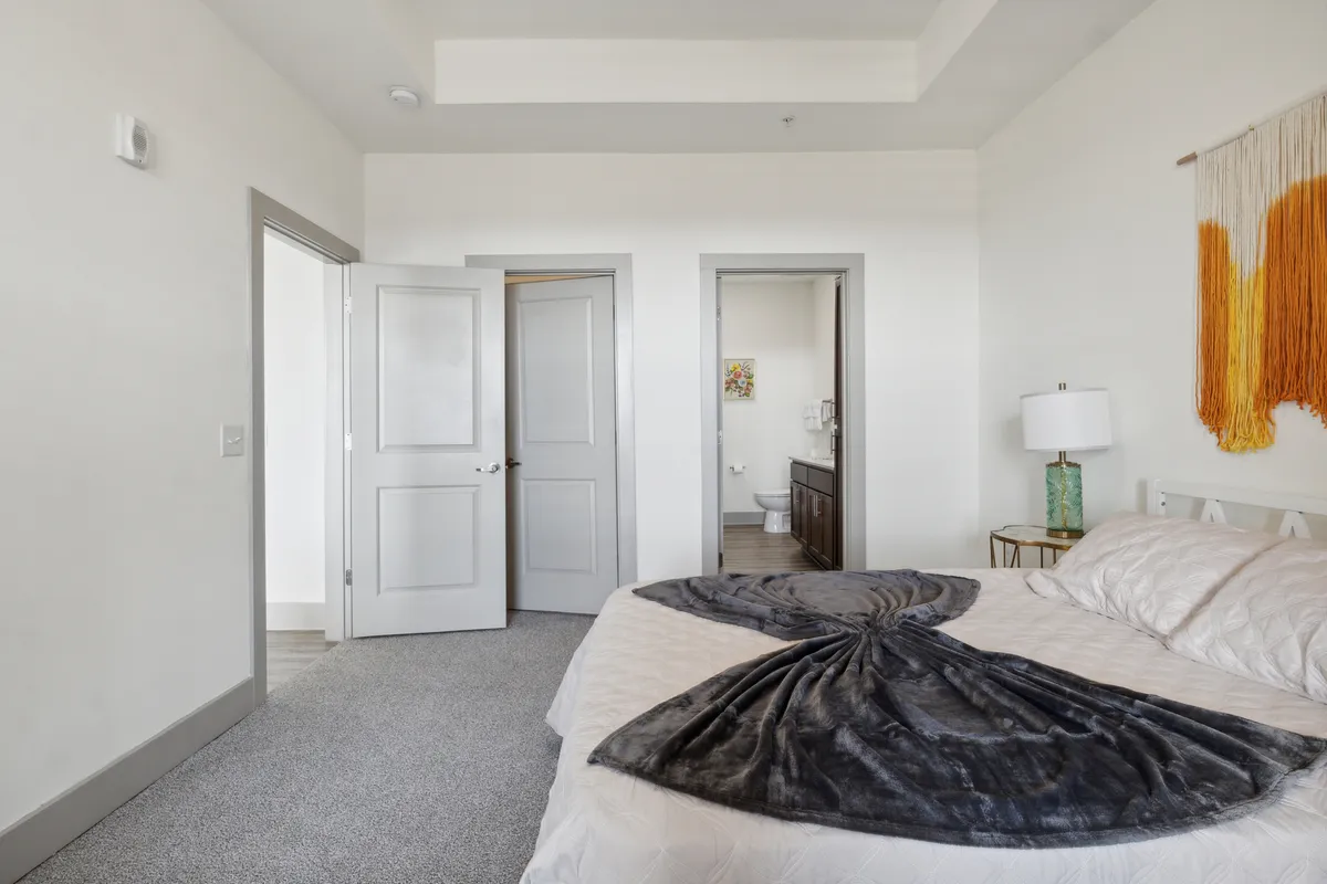 A tranquil bedroom with a decorative blanket on the bed, warm-toned wall decor, and soft natural light streaming in. Doorways hint at an airy, open layout leading to other parts of the home.