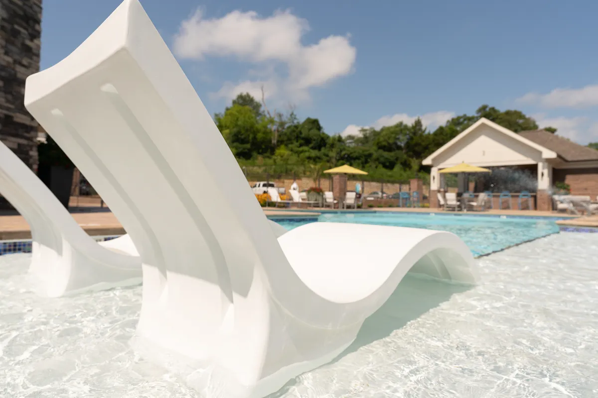 An inviting exterior shot featuring white lounge chairs arrayed along a tranquil pool, with lush greenery and the clubhouse visible in the background. The calm water and serene atmosphere evoke leisurely moments and gatherings in shared outdoor spaces.