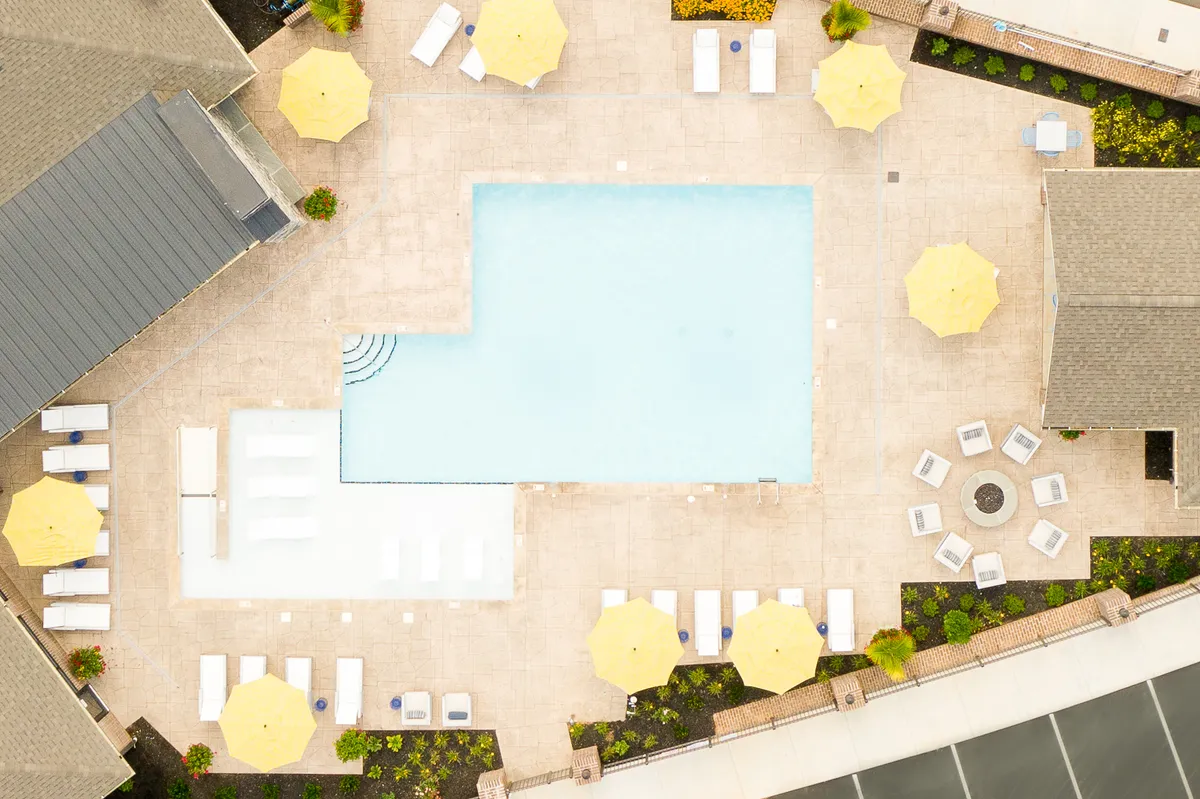 A vibrant exterior view of a community pool area, showcasing sparkling blue water, yellow umbrellas, lounge chairs, and a cozy seating area around a fire pit. The surroundings suggest a cheerful, inviting space ideal for family fun and community gatherings.