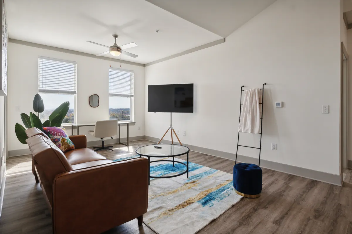 A spacious living area with a modern sofa, coffee table, large TV, and a desk with a chair, all bathed in abundant natural light from expansive windows. The room is decorated in a comfortable, contemporary style, providing both a space to relax and a functional area for work or casual conversation.
