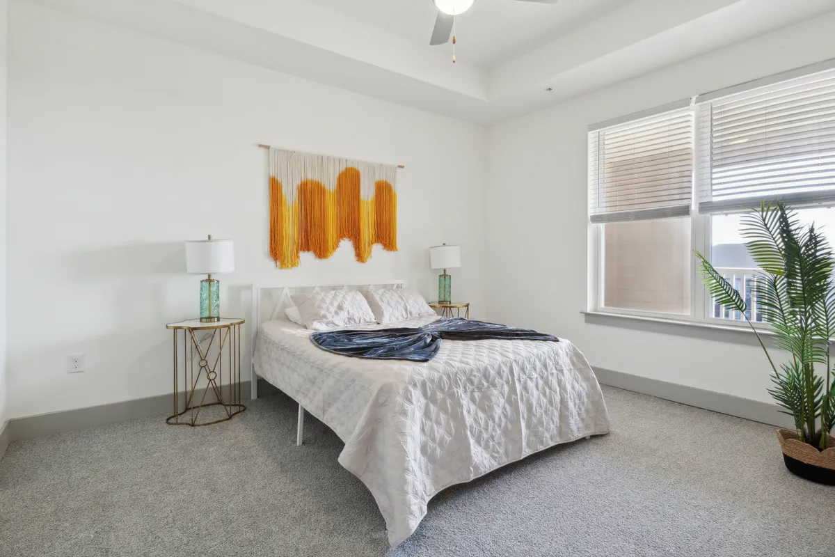 A serene bedroom featuring a bed with a textured quilt, a colorful wall hanging, a green plant, and decorative lamps. Sunlight spills through the window, creating a relaxing, calm oasis ideal for restful sleep or quiet reading.