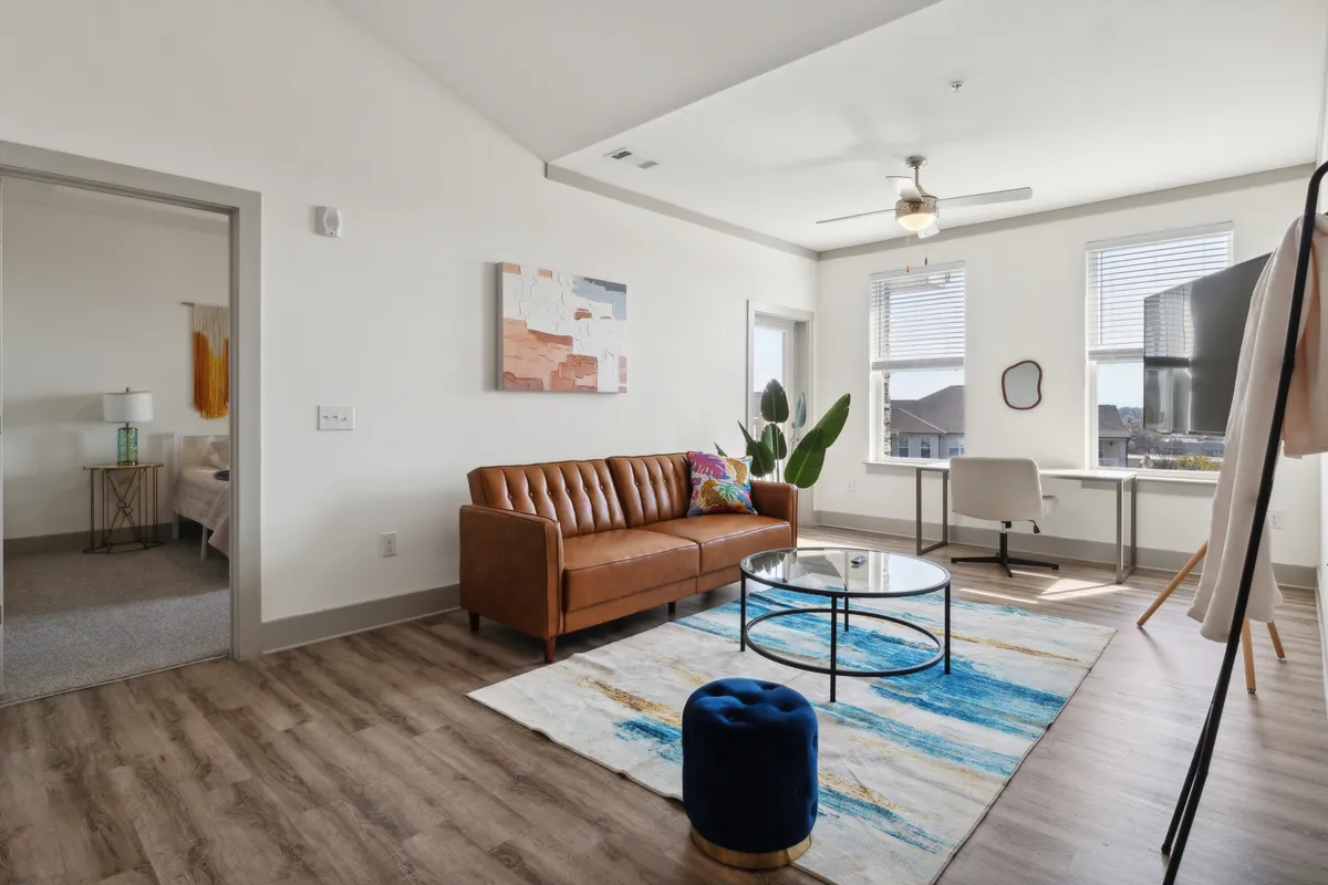 The image showcases a stylish, sunlit living area featuring a plush brown leather sofa, vibrant artwork, a sleek glass coffee table, and a cozy blue ottoman. A modern desk with chair in the corner hints at a versatile space for both relaxation and productivity. The overall ambiance is contemporary, comfortable, and inviting—ideal for unwinding, socializing, or catching up on work.